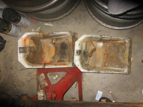 1988 Ford Bronco - Two heavily rusted white metal components are shown resting on a red jack stand, with other automotive parts visible in the background.