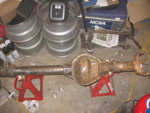 1988 Ford Bronco - A rusty rear axle assembly is supported by jack stands, with spare wheels and a cooler nearby.