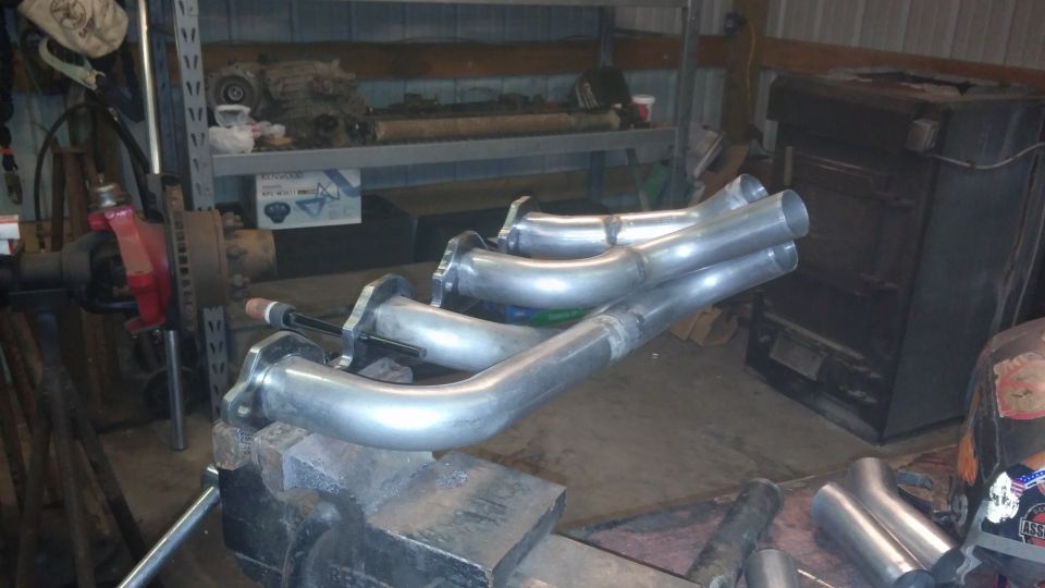 custom exhaust headers clamped in a vise, with a vehicle axle and shelves in the background