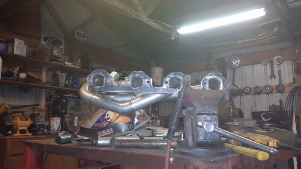 Custom exhaust headers clamped in a vise on a workbench, with welding equipment visible