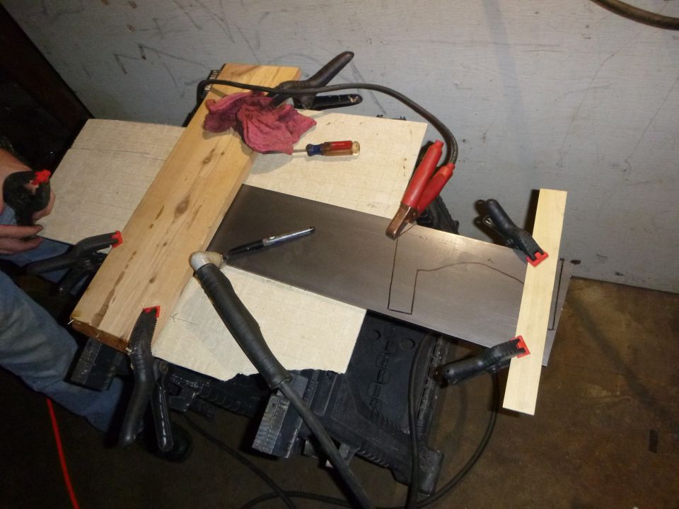 Metal sheet clamped to wood with cutting lines drawn, welding equipment nearby