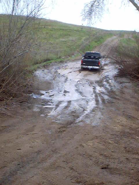 1976 Ford F-100 - A black pickup truck navigates a challenging muddy track with puddles and tire ruts.