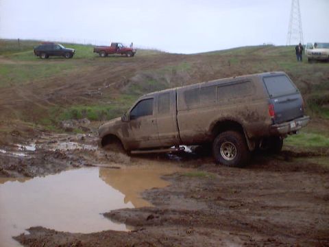 1976 Ford F-100 - A pickup truck is stuck in a mud pit with other vehicles visible on a nearby hill.