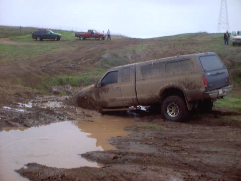1976 Ford F-100 - A pickup truck is stuck deep in the mud, with other vehicles visible on a nearby hill.