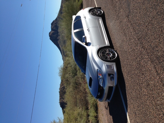 White sedan with a dark hood and aftermarket wheels parked on a paved road with a mountain in the background