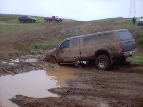 1976 Ford F-100 - A muddy pickup truck with a camper shell is stuck in a muddy rut, with other vehicles in the background.