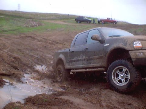 1976 Ford F-100 - Pickup truck navigates a challenging muddy terrain, with other vehicles and individuals in the distance.