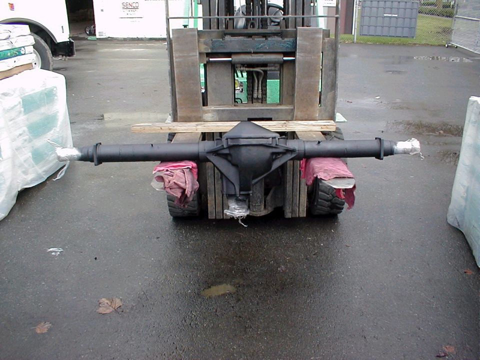 Flat-black rebuilt solid axle housing resting across forklift forks in a wet yard