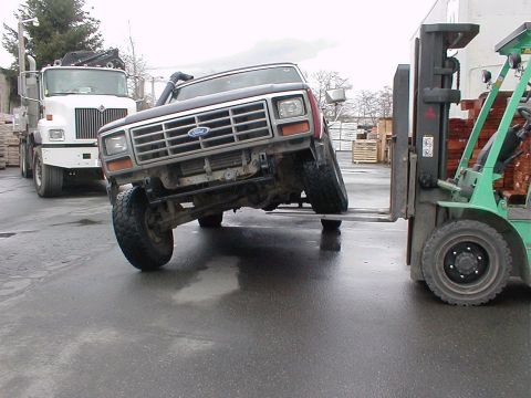 1983 Ford Bronco - A Ford Bronco is being lifted by a forklift, with a large white truck parked behind it.