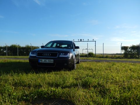 2003 Audi A6 - A dark Audi A6 sedan is parked in a grassy area under a clear blue sky, with a fence and utility structures in the background.