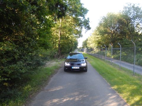 2003 Audi A6 - A black 2003 Audi A6 is seen from the front on a paved path surrounded by trees and a fence.
