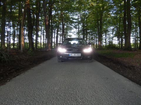 2003 Audi A6 - A black Audi sedan is parked on a gravel road surrounded by trees, its headlights illuminated.