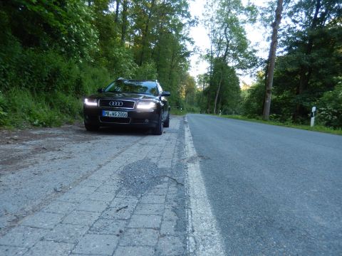 2003 Audi A6 - A dark sedan is parked on a paved shoulder beside a road, with its headlights on.