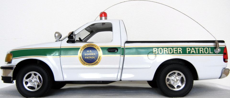 Side view of a white pickup truck with green stripe and 'BORDER PATROL' text. Has a U.S. Border Patrol seal on the door