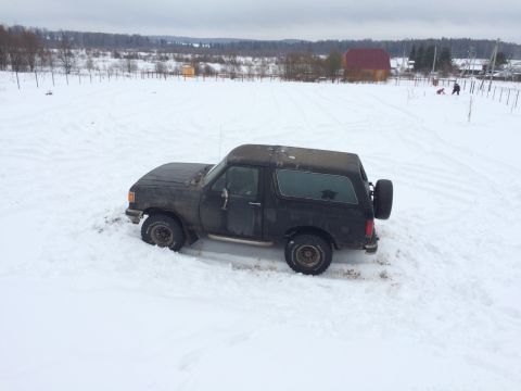 1987 Ford Bronco - A black SUV is stuck in a snowy field with trees and buildings in the background.