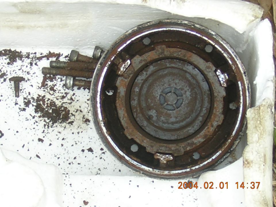 Rusted circular metal part with four bolts and debris on a white surface