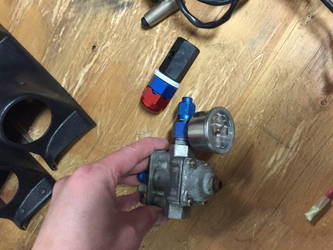 1988 Ford Bronco - A person holds a fuel pressure regulator and gauge, with other car parts and tools scattered on a workbench.