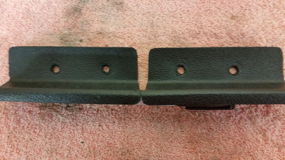 Two black plastic trim pieces with two screw holes each, lying on a pink textured surface. A Supermotors logo is in the lower right
