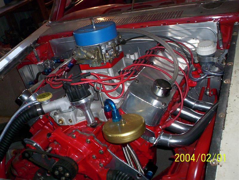 Race-style V8 engine bay with red block, Holley-style carburetor, blue air cleaner base, and aluminum panels