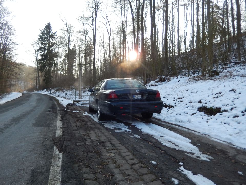Dark sedan parked on a snowy road with trees in the background. The license plate is visible
