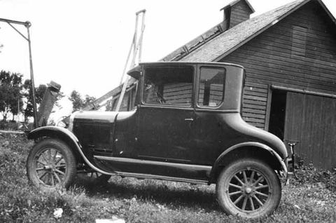 1909 Ford Model T - A dark-colored coupe with wooden spoked wheels is parked on grassy terrain in front of a rustic wooden barn. The vehicle's body style sugge…