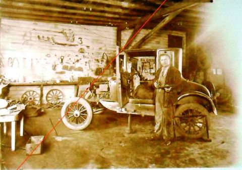 1909 Ford Model T - A man poses with a car in a workshop, surrounded by tools and spare wheels.