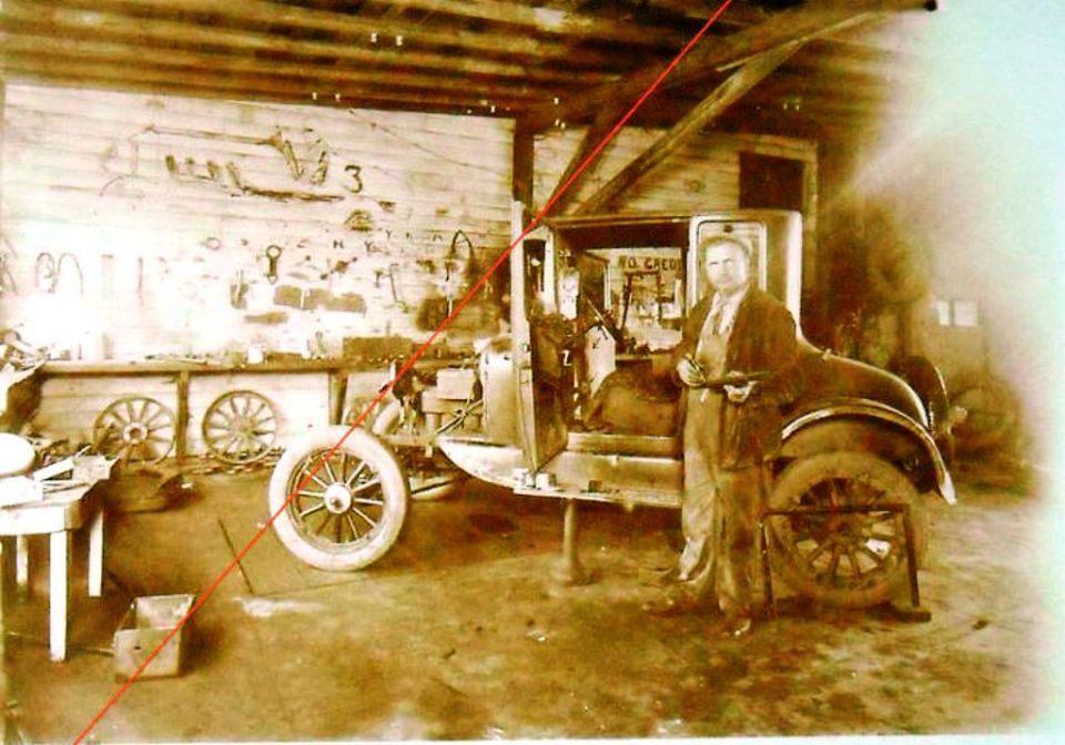 man in workshop with early 1910s car, tools on wall, spare wheels