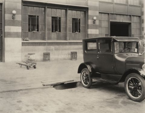 1909 Ford Model T - A dark sedan is parked on concrete in front of a building with large windows. The vehicle appears to be from the mid-1920s.