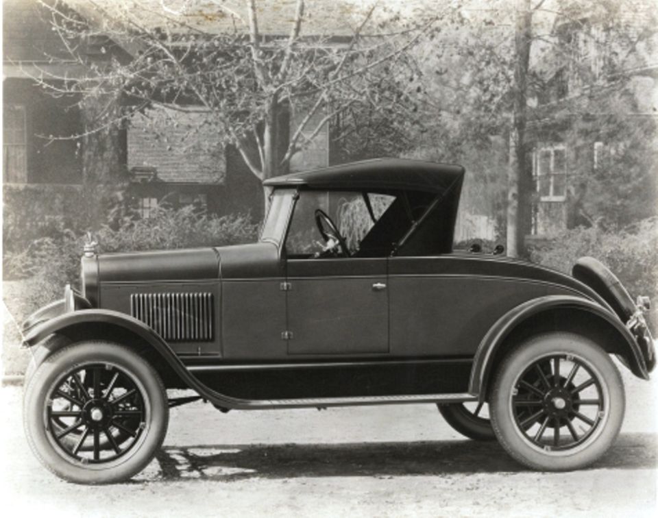 Side view of a dark-colored 1920s convertible coupe with a soft top and spare tire on the rear