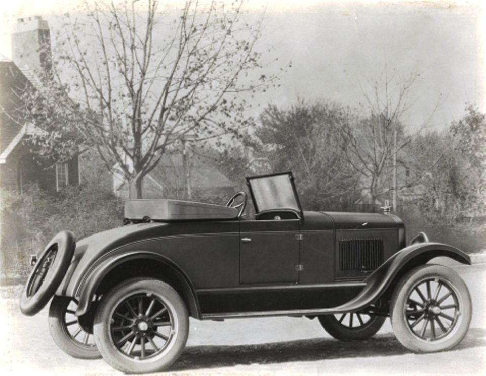 Side view of a dark-colored convertible with a spare tire on the back. It has a tall grille and spoked wheels
