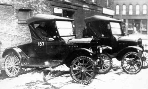 1909 Ford Model T - Two vintage cars are parked on a snowy street in front of buildings.