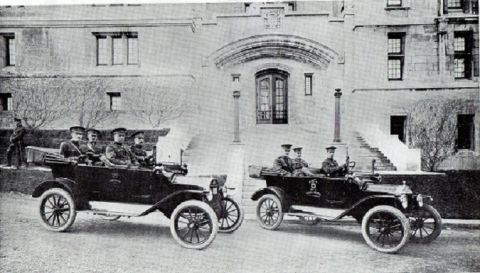 1915 Ford Model T - Two 1915 Ford Model T touring cars, each carrying military personnel, are parked in front of a large building.