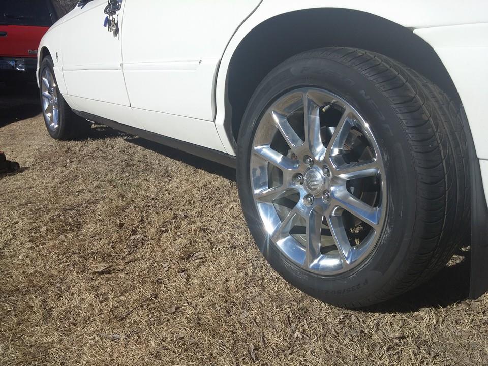 Close-up of a white sedan's rear wheel and side panel. A red vehicle is partially visible in the background