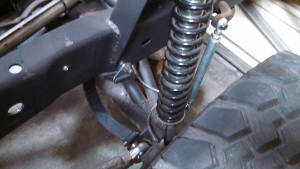 Close-up of a black metal truck frame with a coilover shock absorber and a large tire. The suspension is visible