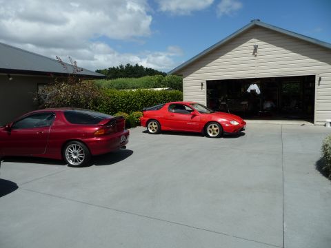 1993 Mazda MX-3 - Two red Mazda MX-3s are parked on a driveway with a garage in the background under a partly cloudy sky.