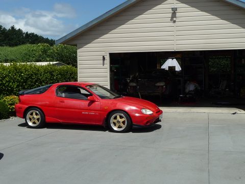 1993 Mazda MX-3 - A red 1993 Mazda MX-3 coupe is parked in front of an open garage on a sunny day.