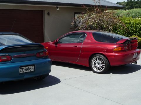 1993 Mazda MX-3 - A red 1993 Mazda MX-3 is parked next to a blue car, showing its rear quarter panel and alloy wheel.
