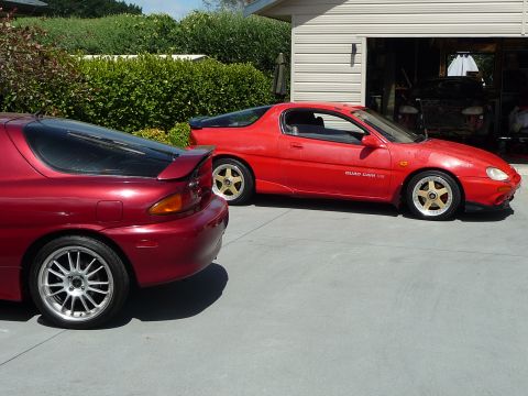 1993 Mazda MX-3 - Two red Mazda MX-3 coupes are parked outside a garage. The car on the right displays a 'QUAD CAM V6' badge.
