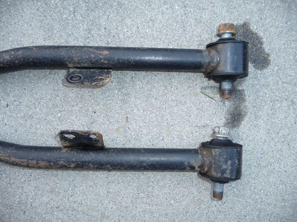 Two black metal suspension control arms with rubber bushings and bolts, resting on concrete