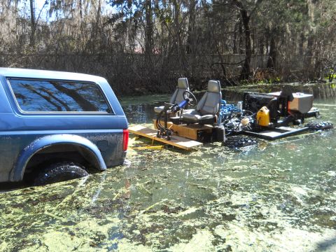 1992 Ford Bronco - A blue 1992 Ford Bronco is partially submerged in a swamp, adjacent to a homemade boat with two seats and an engine.