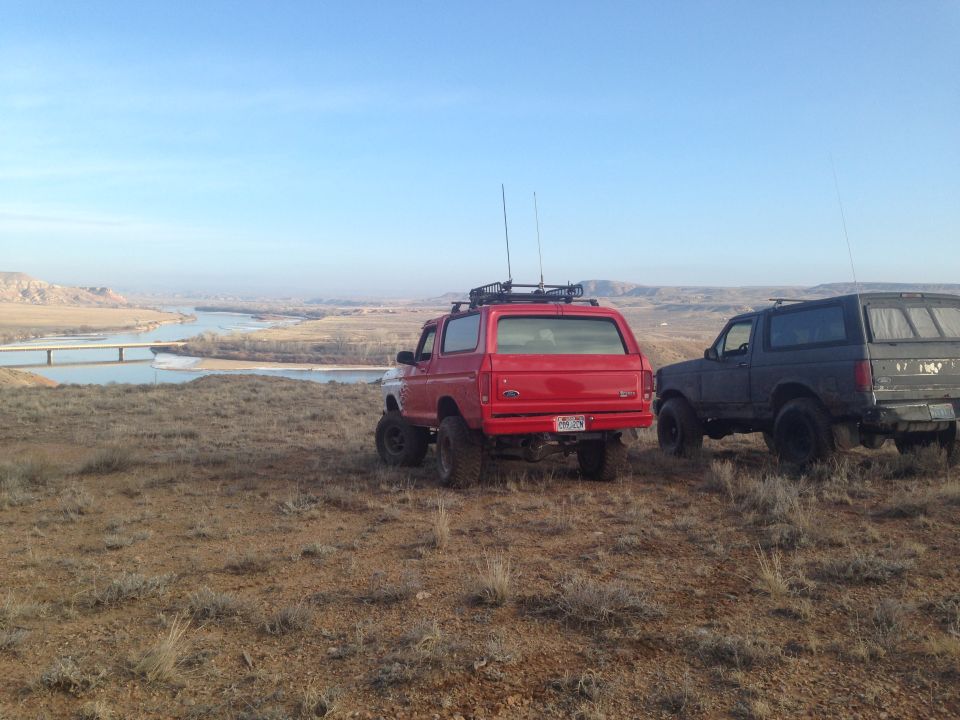 Red 1979 Ford Bronco and a dark SUV parked on a hill with a river in the background