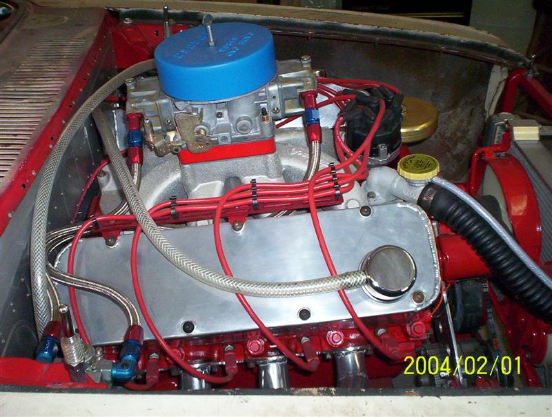 Red-painted carbureted V8 with blue air cleaner, aluminum valve covers, and braided AN lines in an engine bay