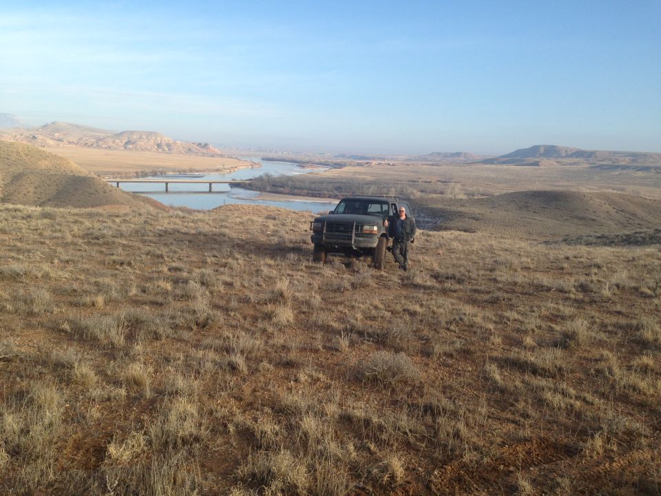 Dark pickup truck on grassy hill with a person, river and bridge in background