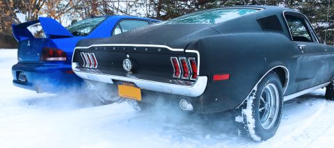 1968 Ford Mustang - A black 1967 Ford Mustang is seen from the rear in a snowy environment, with a blue vehicle behind it.