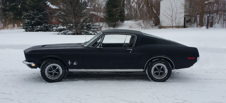 Side view of a black coupe with a fastback roofline parked in the snow. It has chrome trim and five-spoke wheels