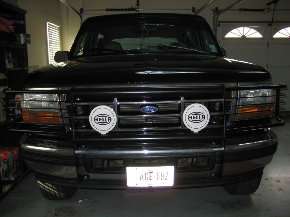 Front of black Ford Bronco in garage with brush guard and Hella light covers, plate AGE 692