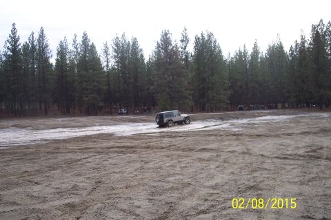 1990 Ford Bronco - A silver Jeep Wrangler navigates a muddy track, with other vehicles and a line of trees visible in the distance.