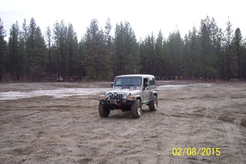 1990 Ford Bronco - A silver Jeep Wrangler sits in a dirt lot with a forest behind it.