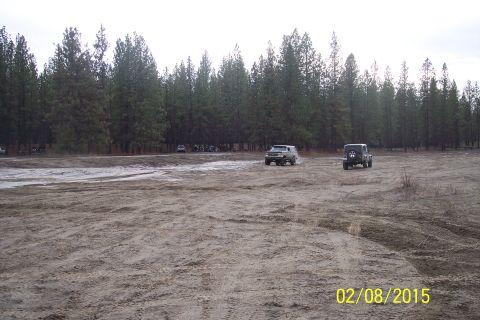 1990 Ford Bronco - Two SUVs navigate a dirt field with a dense forest in the background.