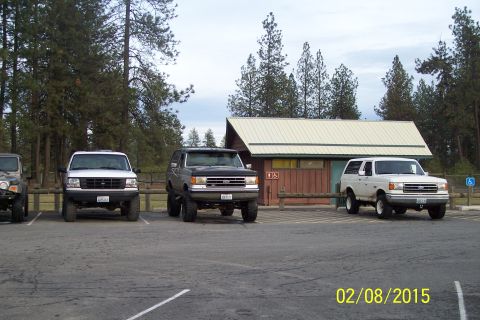 1990 Ford Bronco - Several Ford Broncos are parked in a lot next to a building with a light-colored roof.